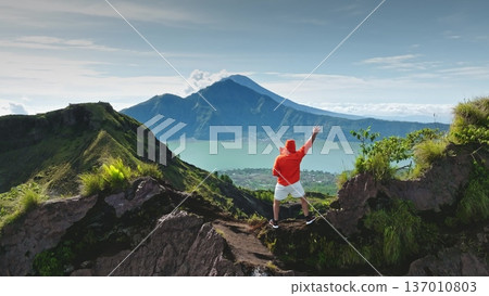 Man standing on Mount Batur ridge in Bali, observing the caldera lake and Mount Agung in the background, celebrating the successful summit and enjoying the rewarding challenge of the adventure Man standing on Mount Batur ridge in Bali, observing the caldera lake and Mount Agung in the background, celebrating the successful summit and enjoying the rewarding challenge of the adventure 137010803