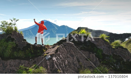 Tourist man celebrate completing challenge hike to the summit of Mount Batur, enjoy personal achievement and overlookinf the stunning green volcanic landscape with Lake Batur in the background Tourist man celebrate completing challenge hike to the summit of Mount Batur, enjoy personal achievement and overlookinf the stunning green volcanic landscape with Lake Batur in the background 137010804