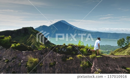 Man runs along Mount Batur volcanic ridge, embracing challenge with Lake Batur and peaks, bright scenic Bali morning. Active sports, travel, hiking outdoor. Zoom out aerial flight panorama flight 137010916