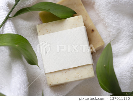 Mockup of beige solid soap bar with unbranded paper label near green leaves on white bath towels 137011120