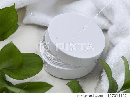 Opened cosmetic cream jar with a blank lid on stone near white bath towel and green leaves, mockup 137011122