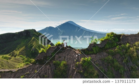 Man enjoying a challenging hike along the rugged ridge of Mount Batur, experiencing adventure and freedom with the breathtaking volcanic landscape and Lake Batur in the background on a bright day 137011173