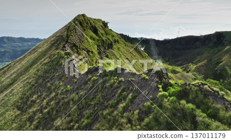 Man running rugged, green covered hills ridge crater rim of Mount Batur volcano, caldera and steaming vents in Bali, Indonesia. Active sports, travel, hiking outdoor. Aerial drone flight panorama 137011179