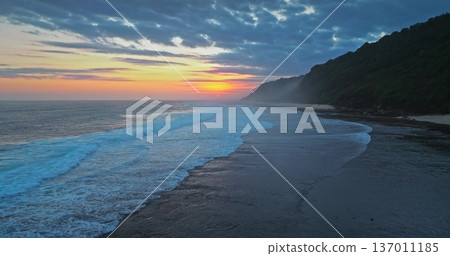 Sunset Bali island coastline with gentle ocean waves rolling sandy beach shore, with orange sun dipping near the horizon, bright colorful clouds in sky, and a lush green cliffside. Aerial drone flight Sunset Bali island coastline with gentle ocean waves rolling sandy beach shore, with orange sun dipping near the horizon, bright colorful clouds in sky, and a lush green cliffside. Aerial drone flight 137011185
