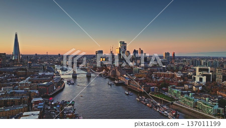 Aerial panorama of London skyline at sunset, with Tower Bridge crossing the River Thames and city landmarks bathed in warm autumn light during dusk. Drone flight over modern cityscape perspective Aerial panorama of London skyline at sunset, with Tower Bridge crossing the River Thames and city landmarks bathed in warm autumn light during dusk. Drone flight over modern cityscape perspective 137011199