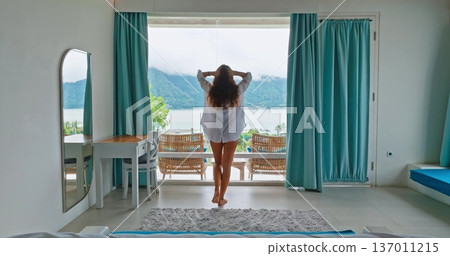 Beautiful young woman in white shirt walking bright hotel room, relax and enjoy serene morning landscape lake view from a wide window, relaxation, travel, and vacation lifestyle in Bali. Back view Beautiful young woman in white shirt walking bright hotel room, relax and enjoy serene morning landscape lake view from a wide window, relaxation, travel, and vacation lifestyle in Bali. Back view 137011215