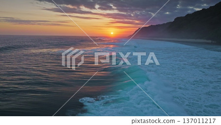 Golden sunset over Indian Ocean coastline, bright colorful clouds sky reflects on vast water surface, powerful waves break tropical Bali island coast with lush green cliffs in background. Aerial shot Golden sunset over Indian Ocean coastline, bright colorful clouds sky reflects on vast water surface, powerful waves break tropical Bali island coast with lush green cliffs in background. Aerial shot 137011217