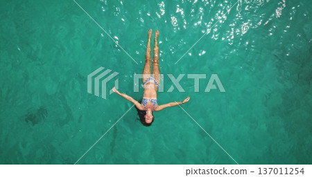 Beautiful young woman relax and enjoy warm, clear sea turquoise water, floating effortlessly on her back, feeling freedom and tranquility during a tropical summer vacation. Aerial top down view Beautiful young woman relax and enjoy warm, clear sea turquoise water, floating effortlessly on her back, feeling freedom and tranquility during a tropical summer vacation. Aerial top down view 137011254