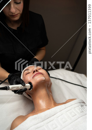 Client relaxing during a skincare procedure. Expert cosmetologist performing anti-aging therapy with a specialized device 137011399