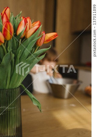 Little girl mixing dough in bowl while mother cooking in background, family baking together at home kitchen Little girl mixing dough in bowl while mother cooking in background, family baking together at home kitchen 137011899
