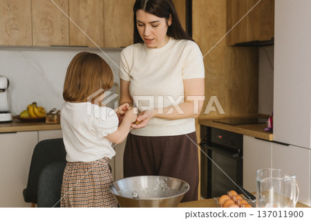Little girl mixing dough in bowl while mother cooking in background, family baking together at home kitchen Little girl mixing dough in bowl while mother cooking in background, family baking together at home kitchen 137011900
