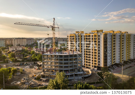 Apartment or office tall building under construction. Brick walls, glass windows, scaffolding and concrete support pillars. Tower crane on bright blue sky copy space background. 137013075