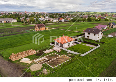 Aerial top view of a private house with attic windows on roof, paved yard with green grass lawn and building site with concrete foundation floor and stacks of yellow bricks for construction.. 137013159