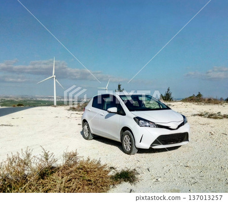 Compact car parked near wind turbines on hill. Sustainable mobility, renewable energy and eco transportation concept Compact car parked near wind turbines on hill. Sustainable mobility, renewable energy and eco transportation concept 137013257