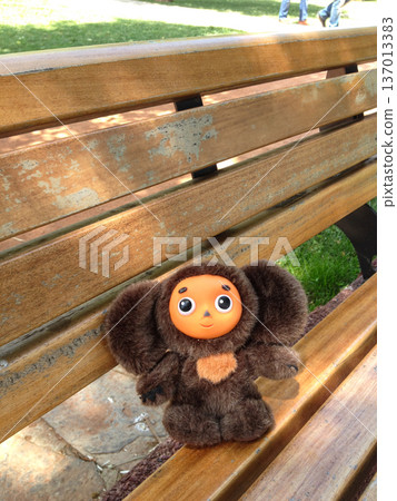 Cheburashka plush toy sitting alone on wooden bench in park. Childhood memory, loneliness, nostalgia and imagination Cheburashka plush toy sitting alone on wooden bench in park. Childhood memory, loneliness, nostalgia and imagination 137013383