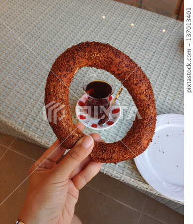 Sesame simit held above tea glass on table. Turkish breakfast ritual, tradition and street food culture concept. Sesame simit held above tea glass on table. Turkish breakfast ritual, tradition and street food culture concept. 137013401