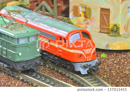 Close-up of two model trains, one red and one green, on miniature railway tracks. Detailed view of a vintage model train set featuring a red M V locomotive alongside a green electric engine on a Close-up of two model trains, one red and one green, on miniature railway tracks. Detailed view of a vintage model train set featuring a red M V locomotive alongside a green electric engine on a 137013455