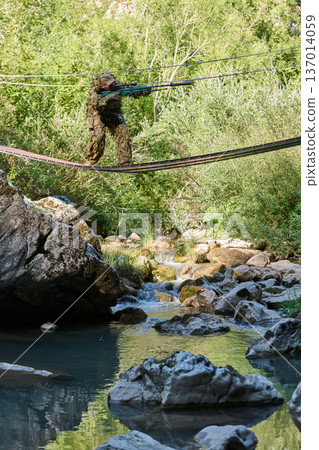 Camouflaged Sniper in Ghillie Suit Crossing Rope Bridge Over Forest Stream During Tactical Training Camouflaged Sniper in Ghillie Suit Crossing Rope Bridge Over Forest Stream During Tactical Training 137014059
