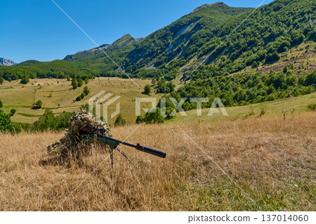 Sniper In Ghillie Suit With Suppressed Rifle Lying In Dry Grass Overlooking Mountain Valley Sniper In Ghillie Suit With Suppressed Rifle Lying In Dry Grass Overlooking Mountain Valley 137014060