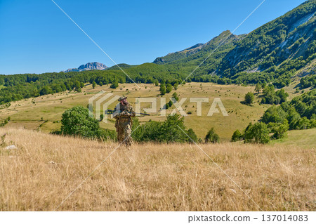 Camouflaged Sniper Standing In Mountain Valley Overlooking Grassy Hills And Forested Slopes Camouflaged Sniper Standing In Mountain Valley Overlooking Grassy Hills And Forested Slopes 137014083