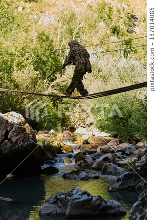 Camouflaged Soldier In Ghillie Suit Crossing Narrow Rope Bridge Over Mountain Stream In Forest Camouflaged Soldier In Ghillie Suit Crossing Narrow Rope Bridge Over Mountain Stream In Forest 137014085