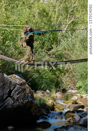 Camouflaged Soldier Sniper Crossing Narrow Suspension Bridge Over Mountain Stream in Forest Training Exercise Camouflaged Soldier Sniper Crossing Narrow Suspension Bridge Over Mountain Stream in Forest Training Exercise 137014092