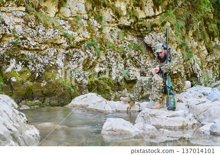 Sniper In Camouflage Sitting On Rocky Riverbank With Rifle In Woodland Ambush 137014101
