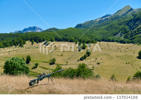 Sniper Rifle on Bipod Overlooking Mountain Meadow Valley With Forested Hills and Clear Blue Sky 137014108