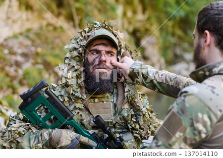 Soldier in Ghillie Suit With Sniper Rifle Receiving Face Paint Adjustment During Field Operation Soldier in Ghillie Suit With Sniper Rifle Receiving Face Paint Adjustment During Field Operation 137014110