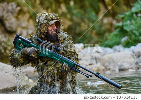Camouflaged Sniper Soldier In Ghillie Suit Wading Through Water With Scoped Rifle In Forest Camouflaged Sniper Soldier In Ghillie Suit Wading Through Water With Scoped Rifle In Forest 137014113