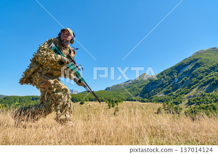 Soldier in Camouflage Ghillie Suit With Rifle on Mountain Surveillance in Sunny Field Soldier in Camouflage Ghillie Suit With Rifle on Mountain Surveillance in Sunny Field 137014124