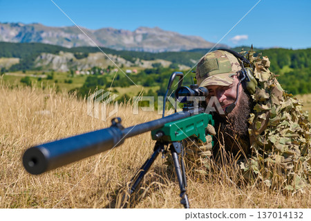 Camouflaged Sniper Soldier Lying In Grass With Scoped Rifle And Suppressor During Field Operation Camouflaged Sniper Soldier Lying In Grass With Scoped Rifle And Suppressor During Field Operation 137014132
