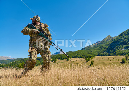 Camouflaged Sniper Soldier With Rifle Patrolling Mountain Field Under Clear Blue Sky 137014152