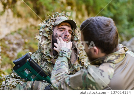 Soldier Applying Camouflage Face Paint During Field Preparation With Ghillie Suit And Rifle 137014154