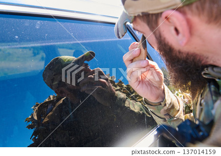 Soldier In Camouflage Inspecting Vehicle Window Reflection During Tactical Field Operation Close-Up Soldier In Camouflage Inspecting Vehicle Window Reflection During Tactical Field Operation Close-Up 137014159