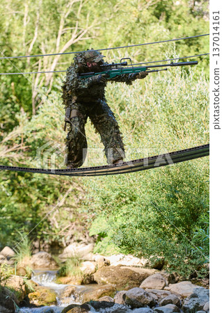 Camouflaged Sniper Soldier Crossing Suspended Rope Bridge Over Stream During Woodland Reconnaissance 137014161