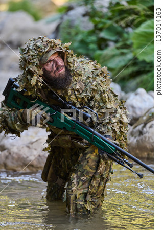 Camouflaged Sniper Soldier In Ghillie Suit Wading Through Stream With Precision Rifle During Reconnaissance 137014163