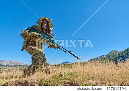 Soldier in Ghillie Suit With Sniper Rifle Moving Through Grass During Mountain Training 137014194
