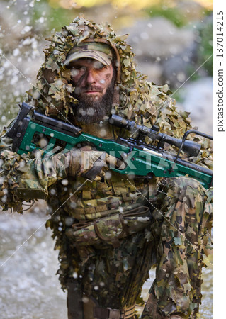 Camouflaged Sniper Soldier In Ghillie Suit With Scoped Rifle Kneeling In Woodland Stream 137014215