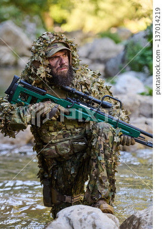 Camouflaged Soldier Sniper In Ghillie Suit With Rifle Kneeling In Rocky River Environment 137014219