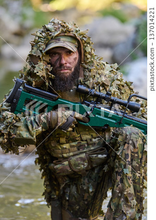 Camouflaged Soldier Sniper In Ghillie Suit With Scoped Rifle Kneeling In Woodland Stream 137014221