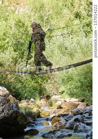 Camouflaged Soldier in Ghillie Suit Crossing Suspended Rope Bridge Over Mountain Stream Camouflaged Soldier in Ghillie Suit Crossing Suspended Rope Bridge Over Mountain Stream 137014222