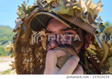 Camouflaged Soldier With Face Paint Wearing Ghillie Hood Applying Camouflage Makeup Outdoors 137014234