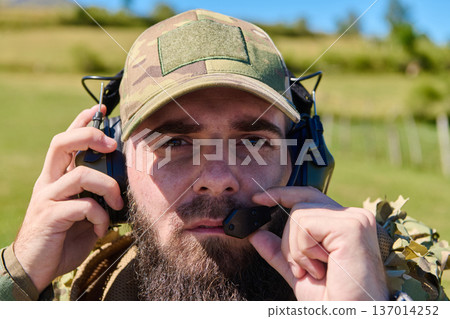 Close-Up of Soldier Wearing Tactical Headset Communicating in Field With Camouflage Cap and Beard 137014252