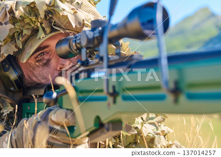 Sniper Soldier in Ghillie Suit Aiming Precision Rifle Through Scope in Sunlit Field 137014257