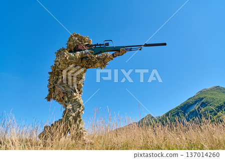 Sniper Soldier in Ghillie Camouflage Aiming a Rifle on Mountain Terrain Under Blue Sky 137014260