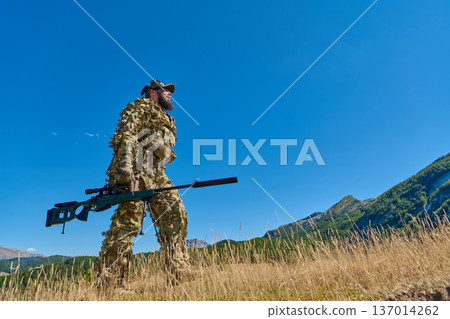 Camouflaged Sniper In Ghillie Suit Walking With Sniper Rifle In Mountain Field Camouflaged Sniper In Ghillie Suit Walking With Sniper Rifle In Mountain Field 137014262