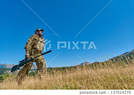 Soldier In Camouflage Ghillie Suit Walking Through Tall Grass With Sniper Rifle In Mountain Landscape 137014270