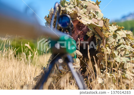 Camouflaged Sniper Soldier In Ghillie Suit Aiming Rifle From Dry Grass During Daytime Training 137014283