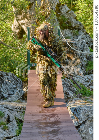 Bearded Soldier In Ghillie Suit Crossing Forest Bridge With Sniper Rifle During Sunny Recon Mission 137014290